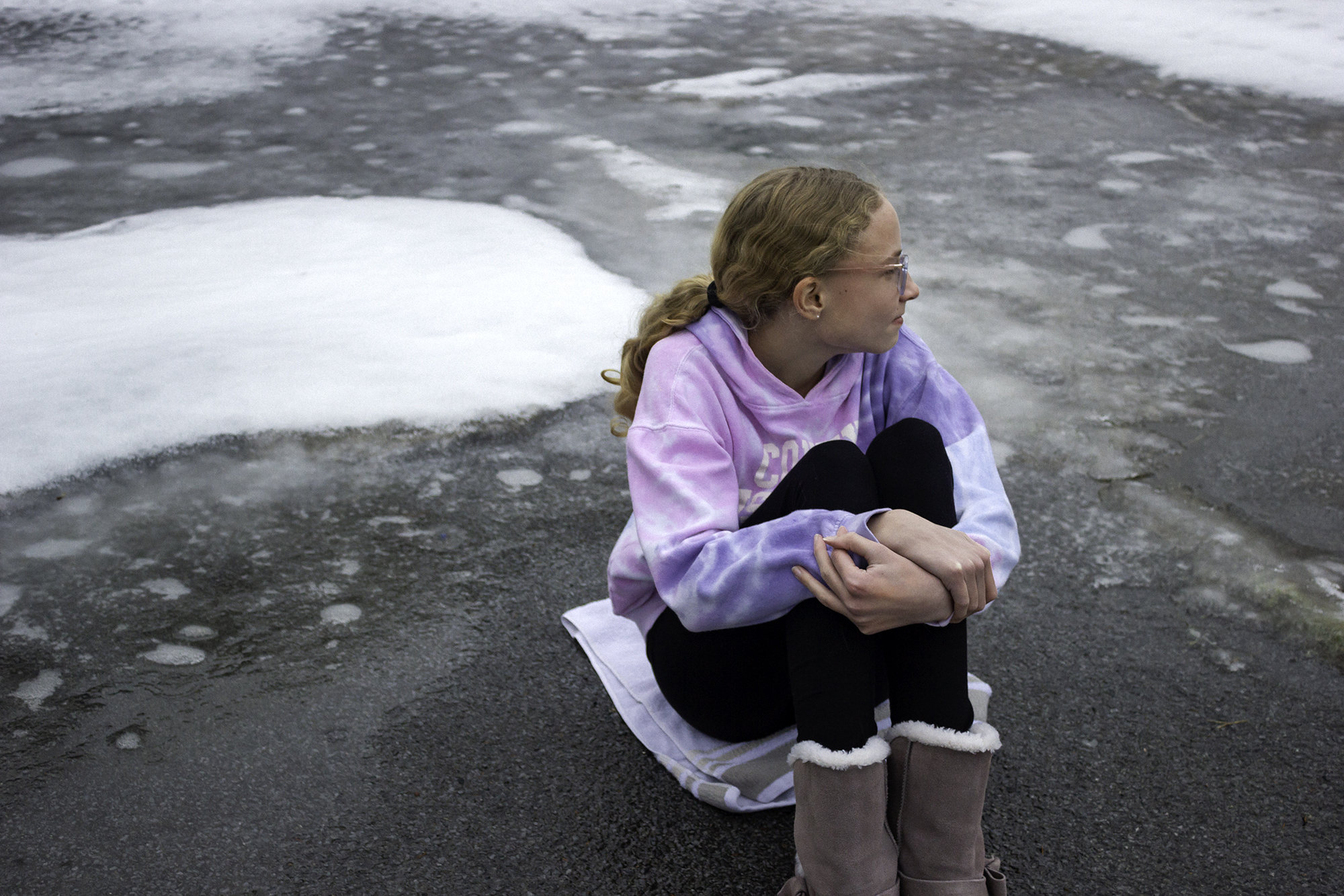 Picture of a young woman sitting on a towel in the middle of an icy and snowy driveway.